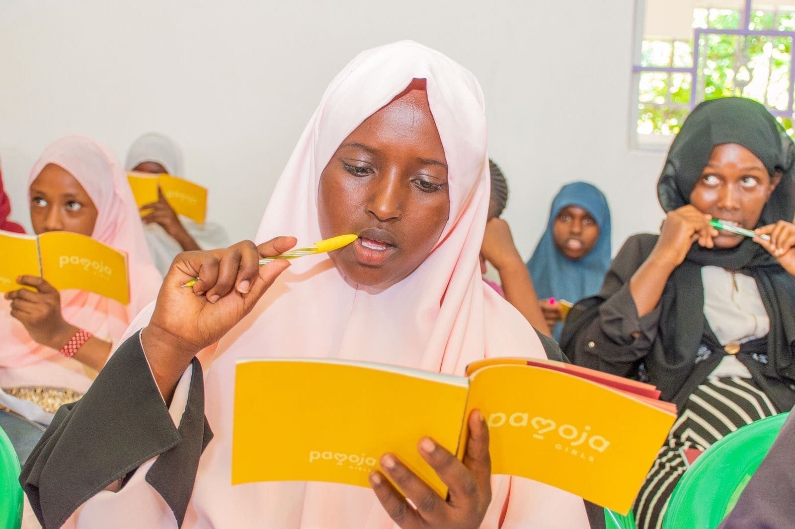 A girl in a pink hijab reads the Girls Book, pen in hand