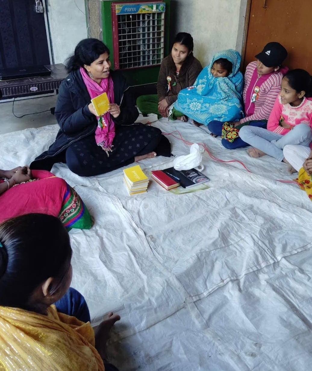 Mona from Sahas holds up a Pamoja Girls Book while girls sit with her in a circle on the floor (Sahas · Atrauli · January 2025)