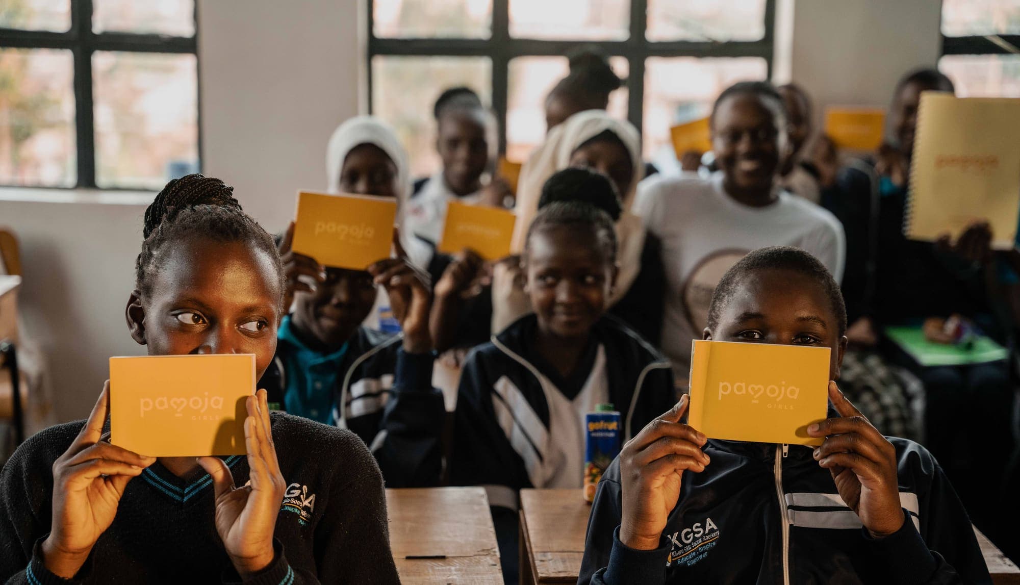 Girls in school uniform hold up their yellow Pamoja Girls Books in a classroom